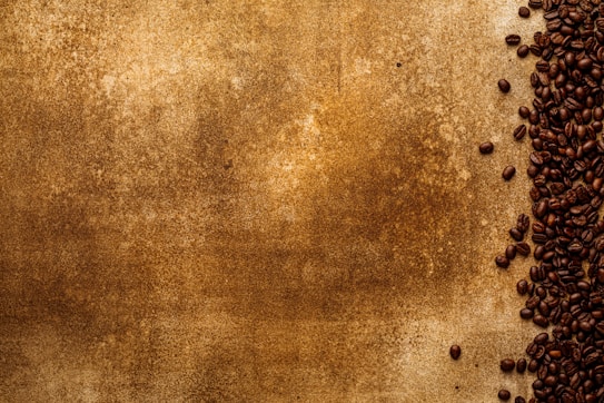 A textured brown background covers most of the image with a collection of dark roasted coffee beans scattered along the right side. The beans are densely packed, creating a contrast between the smooth texture of the beans and the rougher, mottled surface of the background.