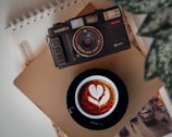 A side view of an UrbanLens camera on a wooden table beside a notebook and coffee cup, capturing a moment of creativity.