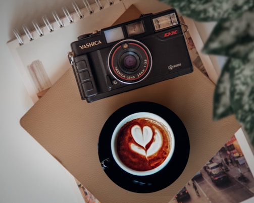 A vintage camera is placed on a beige notebook, with a cup of cappuccino featuring latte art in the shape of a heart nearby. The scene is complemented by a leafy green plant in the corner and a background glimpse of an urban landscape photo.