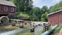 A rustic watermill set in a lush, green environment. The aged red wooden buildings have a classic charm with weathered windows. A wooden waterwheel turns in the flowing stream, surrounded by dense trees and natural vegetation.