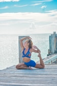 A woman is practicing yoga on a wooden dock by the sea. She is in an advanced pose with one leg bent behind her back, holding her foot with one hand, while the other arm is lifted, touching her head. The background features a calm, expansive ocean with a clear blue sky and some clouds.