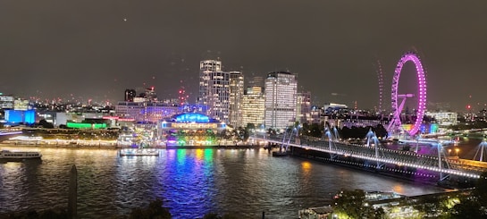 A vibrant cityscape at night with illuminated buildings and a large Ferris wheel glowing in pink. The brightly lit structures reflect on the river, creating a colorful display. A bridge spans the river adorned with lights, and boats are visible on the water.