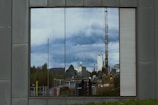A reflective glass window on a metal-clad wall shows a reflection of a cityscape. The reflection includes buildings, including a tall skyscraper and a dome-shaped building, a construction crane, and several people standing in front of the reflection. The sky overhead is cloudy.