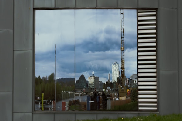 A reflective glass window on a metal-clad wall shows a reflection of a cityscape. The reflection includes buildings, including a tall skyscraper and a dome-shaped building, a construction crane, and several people standing in front of the reflection. The sky overhead is cloudy.