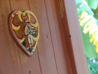 A painted heart-shaped wooden plaque is attached to a vertical brown wooden surface. The plaque features an illustration of a figure in traditional attire, carrying a stringed instrument and wearing a brimmed hat. Bright colors, including yellow and red, accentuate the design, against a background that appears to be surrounded by stylized floral patterns. Part of a leafy green environment is visible at the edge of the image.