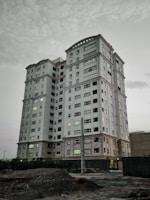 A tall, multi-story residential building with numerous windows, some of which have lights on, indicating occupancy. The building appears to be made of light-colored materials with a modern architectural style. The ground in front of the building is rough and unfinished, suggesting ongoing construction or renovation work. The sky is overcast with clouds, giving a somewhat muted lighting to the scene.