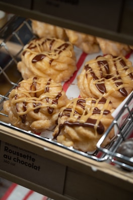 A close-up view of pastries on a metal rack, topped with alternating drizzles of chocolate and caramel. The pastries have a twisted, doughnut-like shape and are placed on a paper lining.