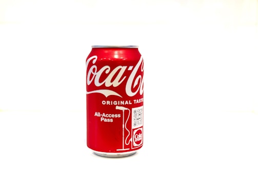 A red aluminum can featuring the Coca-Cola logo in white and promotional elements such as a QR code and the phrase 'All-Access Pass'. The can is placed against a stark white background, emphasizing its colors and design.