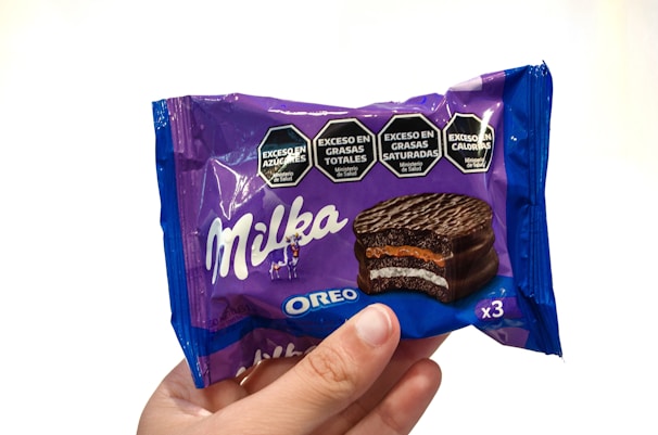A hand holds a Milka Oreo chocolate snack package. The wrapper is predominantly purple with a picture of the chocolate snack, showing layers of chocolate, cream, and an orange filling. The package features hexagonal labels warning about high sugar, total fats, saturated fats, and calories.