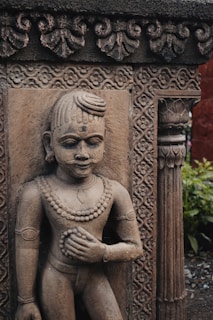 A detailed stone carving of a standing figure adorned with intricate jewelry is embedded in a richly decorated frame with floral and geometric patterns. The figure features stylized hair and a serene expression, exemplifying ancient craftsmanship.
