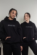 Two individuals are wearing matching black hoodies with 'PARTUM' written on them, posing casually. The background is a plain light wall, creating a minimalist setting.