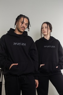 Two individuals are wearing matching black hoodies with 'PARTUM' written on them, posing casually. The background is a plain light wall, creating a minimalist setting.