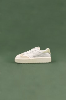 A white sneaker with cream accents and a thick sole is positioned on a smooth, muted green background. The sneaker is designed with a combination of leather and suede materials, with detailed stitching visible along the sides.