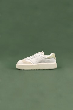 A white sneaker with cream accents and a thick sole is positioned on a smooth, muted green background. The sneaker is designed with a combination of leather and suede materials, with detailed stitching visible along the sides.