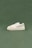 A white sneaker with cream accents and a thick sole is positioned on a smooth, muted green background. The sneaker is designed with a combination of leather and suede materials, with detailed stitching visible along the sides.