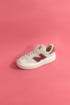 A single sneaker rests on a pink background. The shoe is primarily white with beige and maroon accents, including a large maroon logo on its side. The laces are neatly tied, and the overall style is casual and sporty.