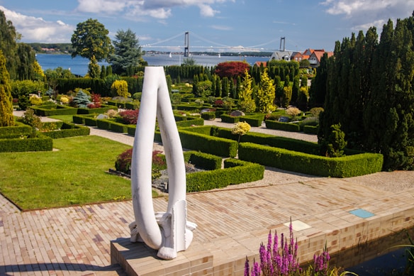 A beautifully manicured garden with neatly trimmed hedges and a variety of trees and shrubs is in the foreground. A prominent modern white sculpture stands on a tiled platform. In the background, a bridge spans a body of water, and houses are visible along the shoreline. The sky is partly cloudy, with patches of blue visible.
