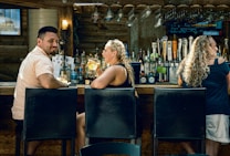 A cozy bar scene with three people seated on high stools. The bar counter is lined with various liquor bottles and a selection of beer taps. Hanging glasses are stored above the counter, and the lighting creates a warm, inviting atmosphere. The individuals appear to be enjoying their time, with one person holding a drink and engaging in conversation.