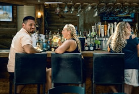 A cozy bar scene with three people seated on high stools. The bar counter is lined with various liquor bottles and a selection of beer taps. Hanging glasses are stored above the counter, and the lighting creates a warm, inviting atmosphere. The individuals appear to be enjoying their time, with one person holding a drink and engaging in conversation.