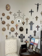 A wall adorned with an assortment of decorative crosses and circular items, with various designs and materials. A beige chair with a floral pattern is positioned nearby, next to a small table holding framed photos, brochures, and a glass dish.