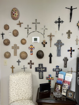 A wall adorned with an assortment of decorative crosses and circular items, with various designs and materials. A beige chair with a floral pattern is positioned nearby, next to a small table holding framed photos, brochures, and a glass dish.