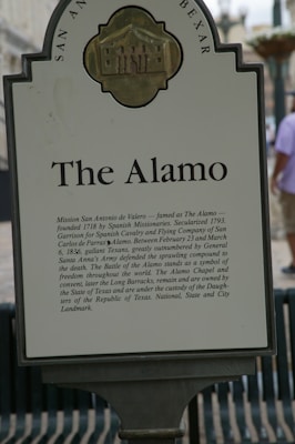 A historical marker detailing information about The Alamo, featuring text that outlines its significance in history as a site defended by Texan forces against Santa Anna's Army. The sign includes a depiction of the mission building at the top and is set against an outdoor backdrop.