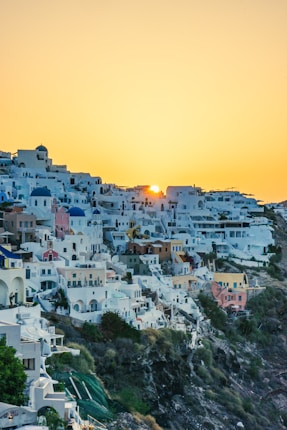 A picturesque hillside village with white-washed buildings and blue-domed roofs, typical of Greek architecture, with a sunset casting a warm glow over the scene.