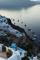 A picturesque coastal landscape with whitewashed buildings built on a hillside overlooking a serene body of water. Several yachts and sailboats are anchored nearby, and a calm atmosphere is accentuated by the soft lighting and clear sky. The cliffside architecture is complemented by pools and outdoor terraces, offering a luxurious and tranquil setting.
