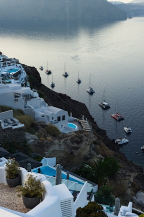 A picturesque coastal landscape with whitewashed buildings built on a hillside overlooking a serene body of water. Several yachts and sailboats are anchored nearby, and a calm atmosphere is accentuated by the soft lighting and clear sky. The cliffside architecture is complemented by pools and outdoor terraces, offering a luxurious and tranquil setting.