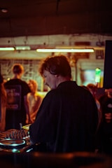 A DJ wearing headphones is performing at a small, intimate club. The setting is dimly lit with warm tones, and a group of people are seen in the background enjoying the music. The DJ is focused on the equipment, which is illuminated by colorful lights.