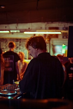 A DJ wearing headphones is performing at a small, intimate club. The setting is dimly lit with warm tones, and a group of people are seen in the background enjoying the music. The DJ is focused on the equipment, which is illuminated by colorful lights.