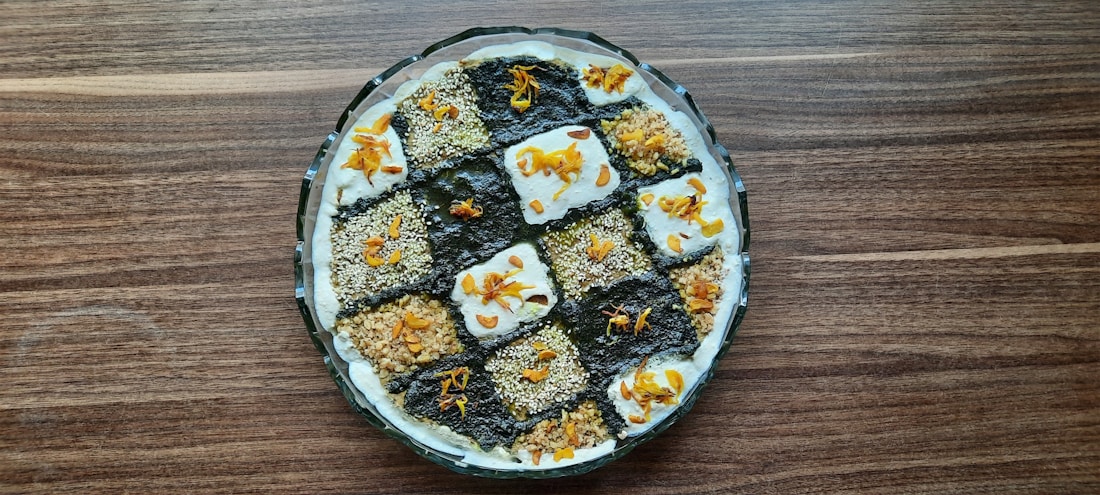 A round dish is filled with a visually appealing dessert featuring a checkerboard pattern. The squares alternate between a dark, chocolate-like texture and a lighter, creamy one, each garnished with nuts and orange-colored toppings. The dessert is presented on a wooden surface, providing a rustic backdrop.