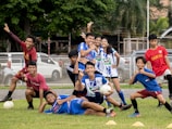 A group of young people in sports uniforms are posing cheerfully on a grassy field. Some are holding a soccer ball, while others are striking playful poses. They seem to be in the midst of a lighthearted moment, possibly after a game or practice session.