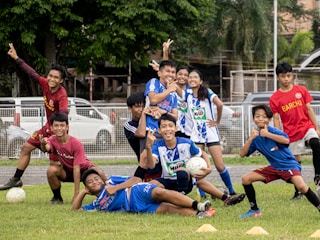 A group of young people in sports uniforms are posing cheerfully on a grassy field. Some are holding a soccer ball, while others are striking playful poses. They seem to be in the midst of a lighthearted moment, possibly after a game or practice session.