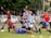 A group of young people in sports uniforms are posing cheerfully on a grassy field. Some are holding a soccer ball, while others are striking playful poses. They seem to be in the midst of a lighthearted moment, possibly after a game or practice session.
