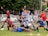 A group of young people in sports uniforms are posing cheerfully on a grassy field. Some are holding a soccer ball, while others are striking playful poses. They seem to be in the midst of a lighthearted moment, possibly after a game or practice session.