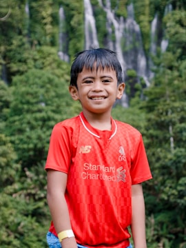 A young boy is standing outdoors, smiling at the camera. He is wearing a bright red sports jersey with a logo and text on it. The background is lush and green with tall waterfalls cascading down a rocky cliff.