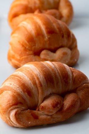 Three golden-brown croissants are lined up diagonally on a white surface. The croissants have a flaky and crisp texture with visible layers. They are well-baked and have a warm, inviting appearance.
