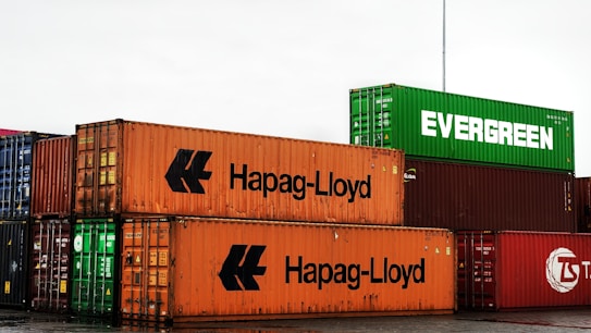 Several shipping containers are stacked in a yard, featuring brands like Hapag-Lloyd and Evergreen. The containers are in various colors including orange and green.