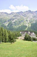 A lush green meadow stretches out in the foreground, bordered by dense forests and set against a backdrop of majestic mountain ranges. Nestled within the trees is a large, multi-story building with a modern architectural design.