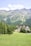 A lush green meadow stretches out in the foreground, bordered by dense forests and set against a backdrop of majestic mountain ranges. Nestled within the trees is a large, multi-story building with a modern architectural design.