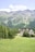 A lush green meadow stretches out in the foreground, bordered by dense forests and set against a backdrop of majestic mountain ranges. Nestled within the trees is a large, multi-story building with a modern architectural design.
