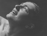 A close-up, monochrome image of a person's face angled upwards, displaying droplets of water on their skin. The expression is one of joy or relief, with eyes partially closed and a slight smile revealing teeth. The lighting emphasizes the texture and contours of the face, giving an intense focus on the expression.