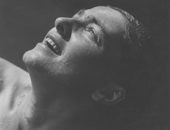 A close-up, monochrome image of a person's face angled upwards, displaying droplets of water on their skin. The expression is one of joy or relief, with eyes partially closed and a slight smile revealing teeth. The lighting emphasizes the texture and contours of the face, giving an intense focus on the expression.