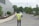 A person wearing a yellow reflective vest stands on a paved road with their hands behind their back. They are facing away from the camera, looking towards a gray building and a few evergreen trees. In the background, another person is walking on the sidewalk, and a vehicle is partially visible.