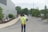 A person wearing a yellow reflective vest stands on a paved road with their hands behind their back. They are facing away from the camera, looking towards a gray building and a few evergreen trees. In the background, another person is walking on the sidewalk, and a vehicle is partially visible.