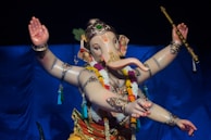 A colorful statue of a deity with an elephant head and multiple arms, adorned with ornate jewelry and floral garlands. The figure holds various objects, including a flute, and is set against a blue background.