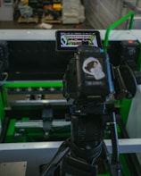 A digital camera is mounted on a tripod, capturing an image of machinery or equipment from a screen. The screen displays a detailed view of the equipment. The setting appears to be industrial with metallic and green components.