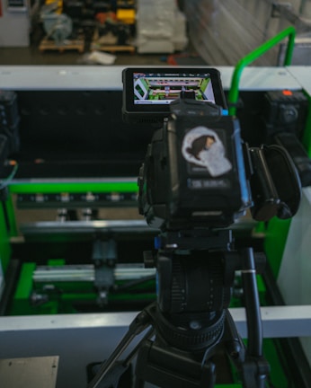 A digital camera is mounted on a tripod, capturing an image of machinery or equipment from a screen. The screen displays a detailed view of the equipment. The setting appears to be industrial with metallic and green components.
