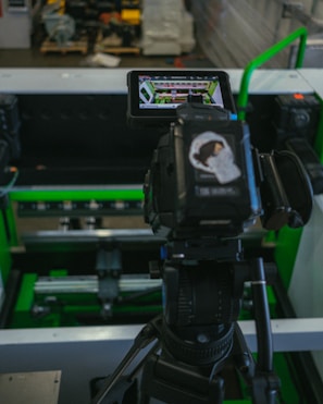 A digital camera is mounted on a tripod, capturing an image of machinery or equipment from a screen. The screen displays a detailed view of the equipment. The setting appears to be industrial with metallic and green components.